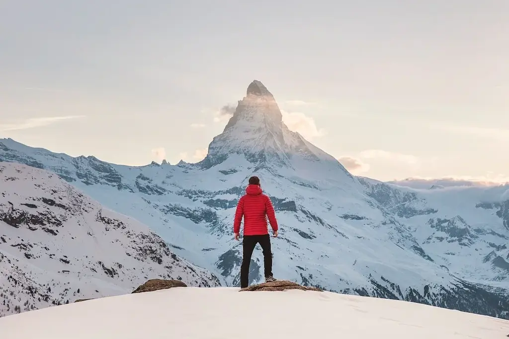 Man in a red jacket standing on a mountain looking at a mountain peek in the distance during winter.