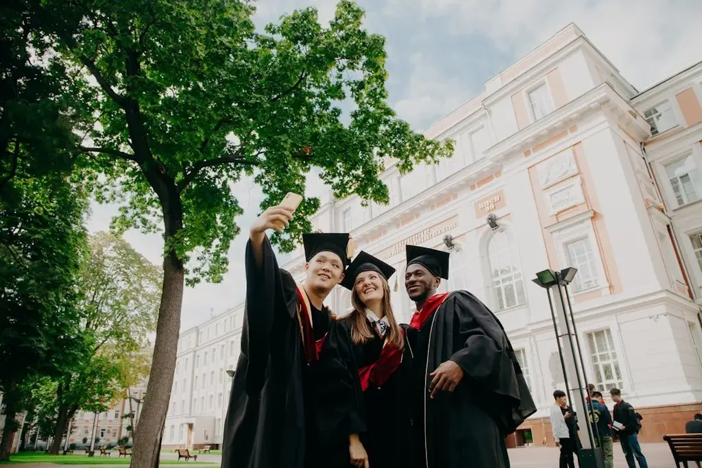Three graduates take a selfie in the graduate gowns with the university in the background